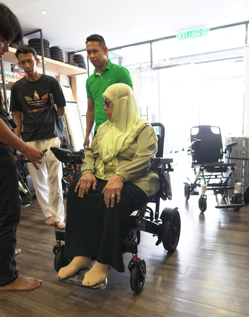 Senior woman testing an electric wheelchair in a Malaysian store with staff. Electric wheelchair test drive.