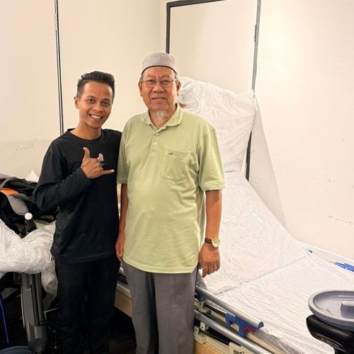 Two men stand near a new hospital bed, one making a shaka sign.