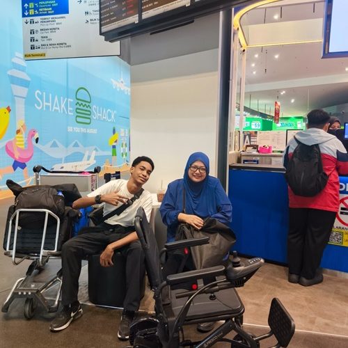 Two travelers with luggage and wheelchairs at an airport, near a Shake Shack.